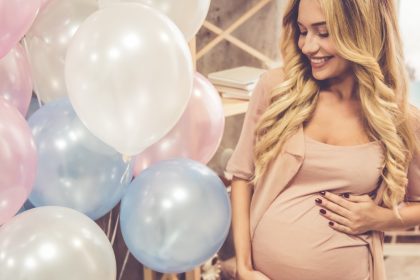 A pregnant woman wearing a beige dress stands next to pink, white, and blue inflated helium balloons.