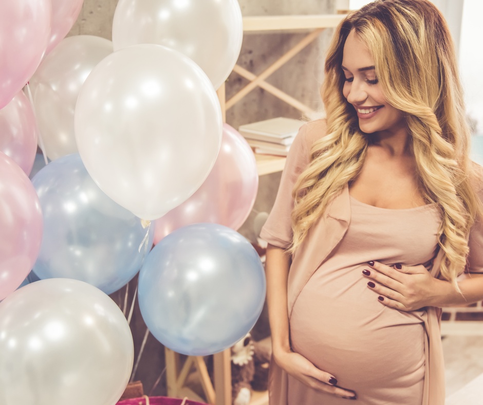 A pregnant woman wearing a beige dress stands next to pink, white, and blue inflated helium balloons.