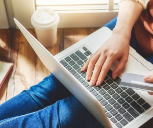 Someone sits on the floor with a laptop open in their lap. They hold a credit card in one hand and type with the other.