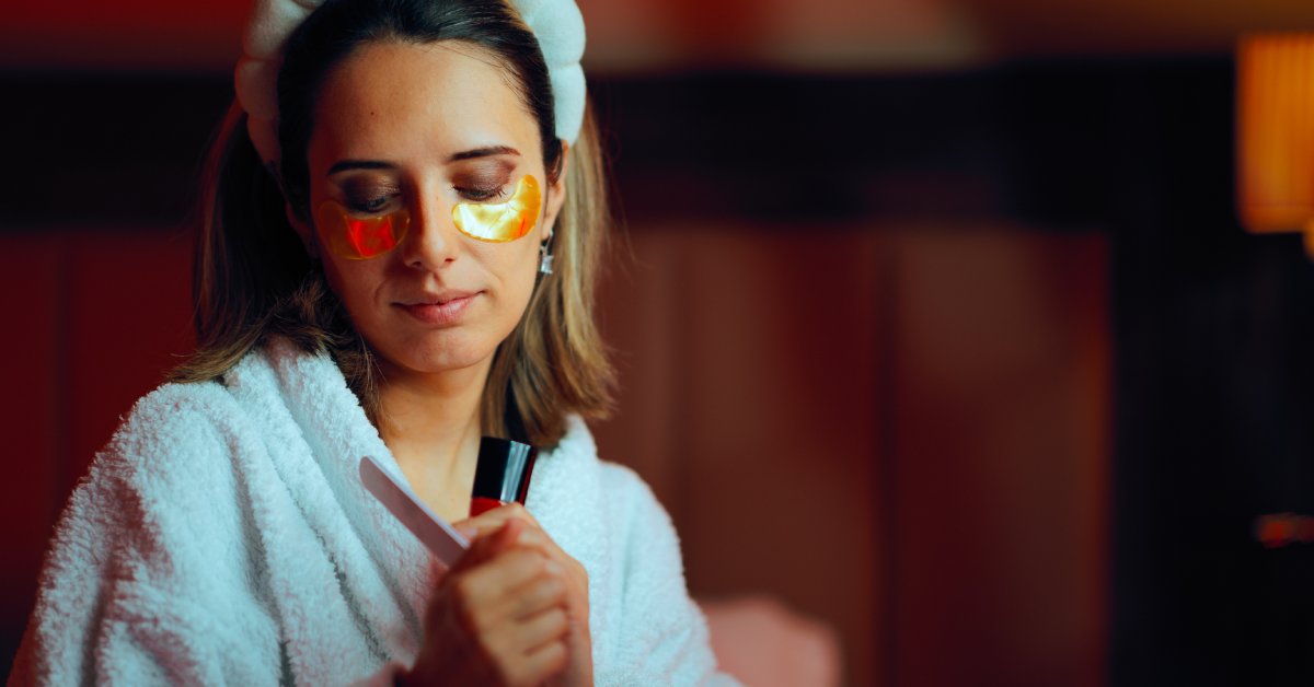 A woman in a white, fluffy robe wears under-eye pads. She looks down at her nails as she files them.