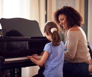 A curly brown haired woman and a little girl in a blue shirt sitting at a piano bench, playing the piano in front of them.
