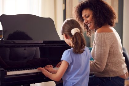 A curly brown haired woman and a little girl in a blue shirt sitting at a piano bench, playing the piano in front of them.