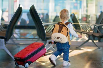 A toddler walking through an airport. They are pulling a red luggage and wearing a backpack in the shape of a lion's head.