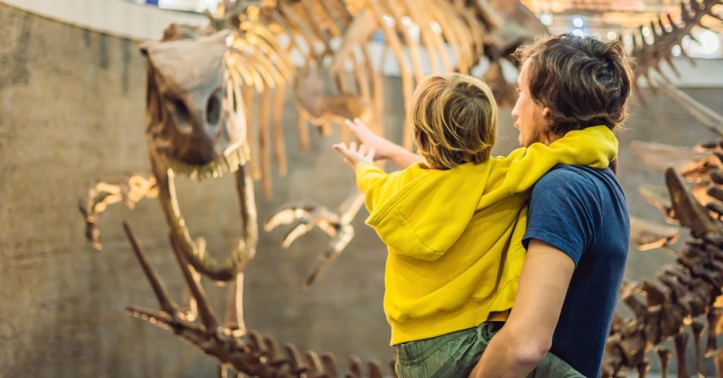 A man inside a museum holding a little boy in a yellow sweatshirt and pointing at a large T-Rex skeleton.