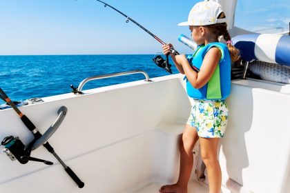 A young girl wearing a hat, shorts, and a life jacket holding a fishing rod while standing on the side of a white boat.