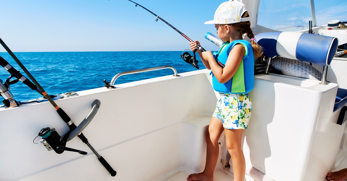 A young girl wearing a hat, shorts, and a life jacket holding a fishing rod while standing on the side of a white boat.