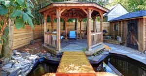 A backyard with an in-ground pond and a bridge leading up to a wooden gazebo with lawn chairs inside.
