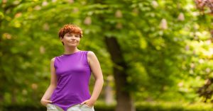 A person with short curly red hair is standing outside in a park. They're wearing a purple sleeveless top and white pants.