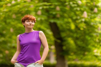 A person with short curly red hair is standing outside in a park. They're wearing a purple sleeveless top and white pants.