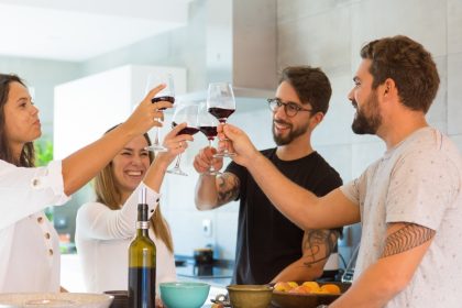 A group of four young adults standing around a kitchen island clink their red wine glasses together.