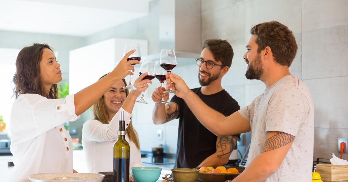 A group of four young adults standing around a kitchen island clink their red wine glasses together.