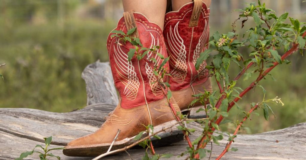 Someone wearing a pair of red and brown leather Western boots while standing on a log in the middle of a green field.