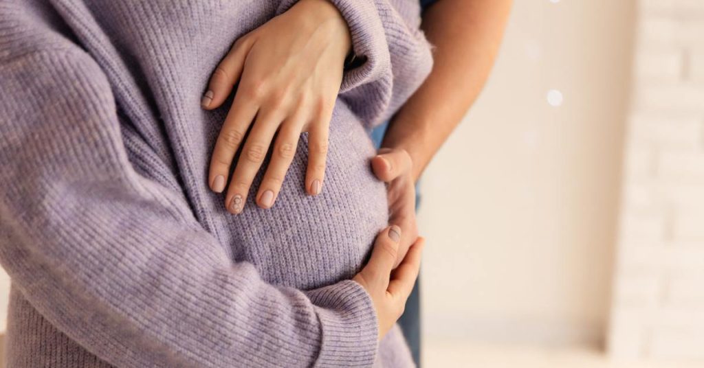 A young family, a pregnant woman wearing a purple sweater, and her partner are hugging and admiring her tummy.