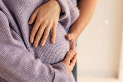 A young family, a pregnant woman wearing a purple sweater, and her partner are hugging and admiring her tummy.