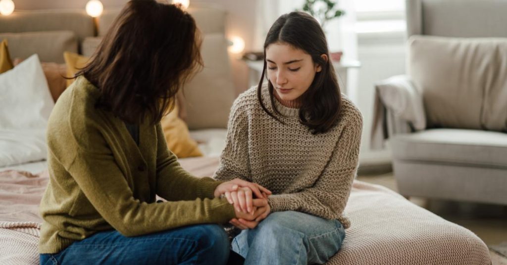 A mother sits beside her teenage daughter on a bed, looking at her. They are holding each other's hands.