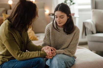 A mother sits beside her teenage daughter on a bed, looking at her. They are holding each other's hands.