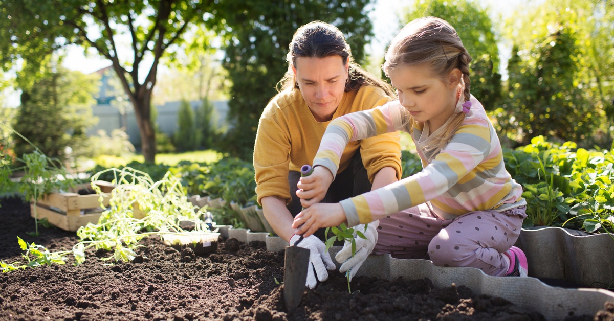 A mom and daughter kneel in a garden as they plant some seedlings. The young daughter holds a trowel as she gardens.