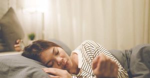A young woman laying on her side with her head and hand on a gray pillow in a well-lit, neutral bedroom space.