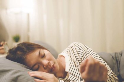 A young woman laying on her side with her head and hand on a gray pillow in a well-lit, neutral bedroom space.