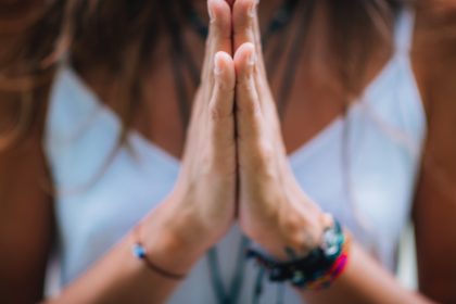 A close-up of a woman pressing her hands together in meditative prayer. She's wearing many bracelets and a white tank top.