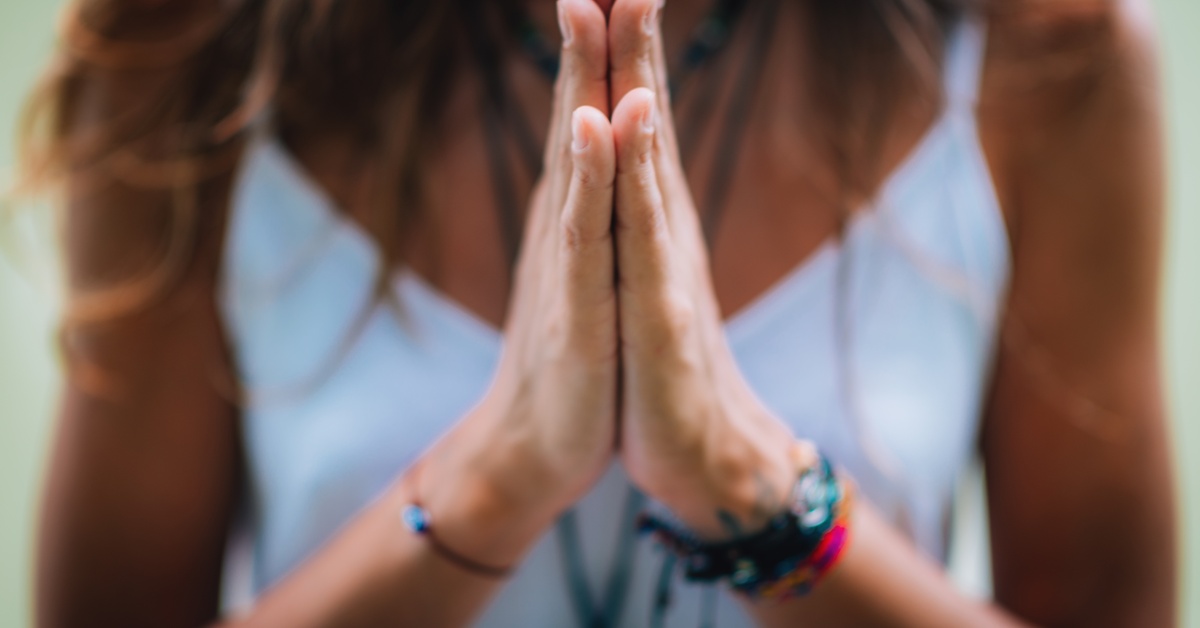 A close-up of a woman pressing her hands together in meditative prayer. She's wearing many bracelets and a white tank top.