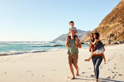 Two parents walk on a beach carrying their two children; one rides on the dad’s shoulders, the other in the mom’s arms.