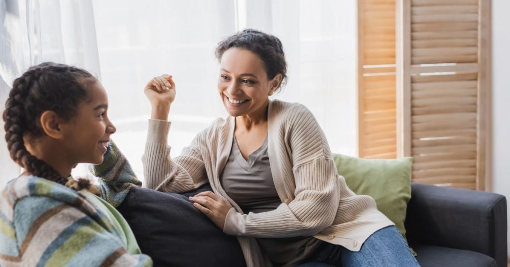 A smiling and laughing woman and her daughter are talking on the couch at home by a window on a sunny day.