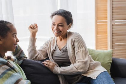 A smiling and laughing woman and her daughter are talking on the couch at home by a window on a sunny day.