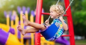 A smiling little boy with blonde hair wears a blue, green, and yellow checkered shirt while on blue swing.