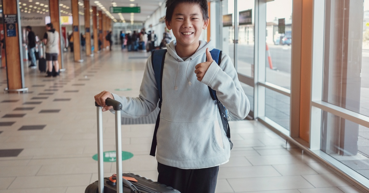 A young boy stands in an airport and gives a thumbs-up gesture. He holds the handle of a rolling suitcase.