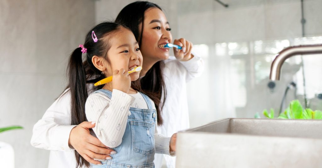 A mom shows her daughter how to brush her teeth as they stand side by side in a bathroom, facing the sink.