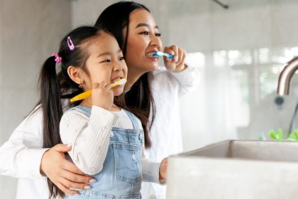 A mom shows her daughter how to brush her teeth as they stand side by side in a bathroom, facing the sink.