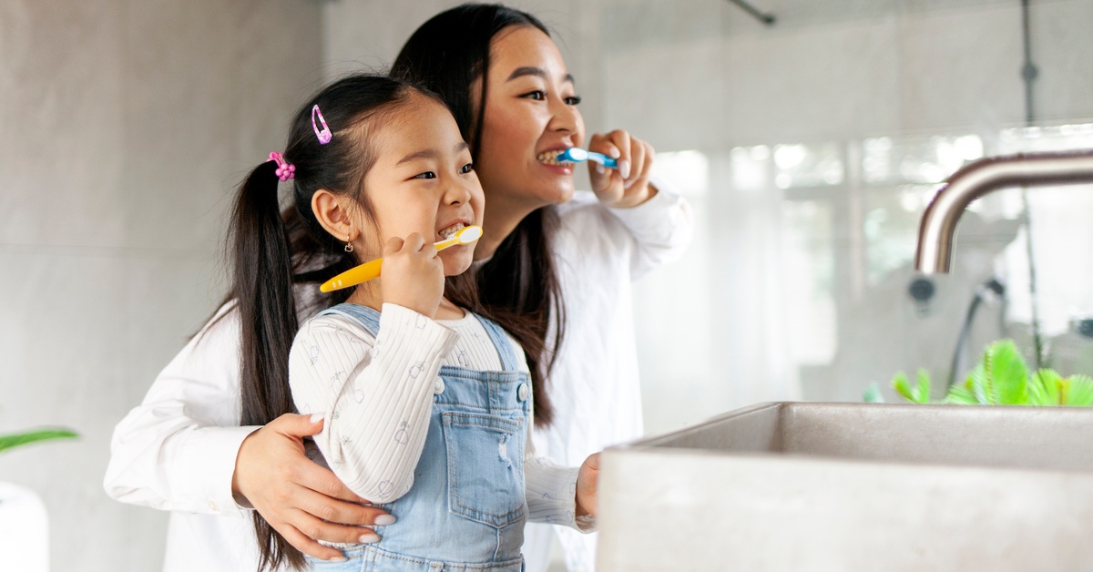 A mom shows her daughter how to brush her teeth as they stand side by side in a bathroom, facing the sink.