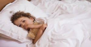 A young child with curly hair in pajamas hugs a teddy bear while lying on a bed with white bedding, gazing off to the side.