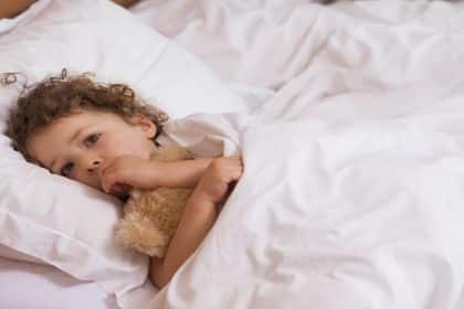 A young child with curly hair in pajamas hugs a teddy bear while lying on a bed with white bedding, gazing off to the side.