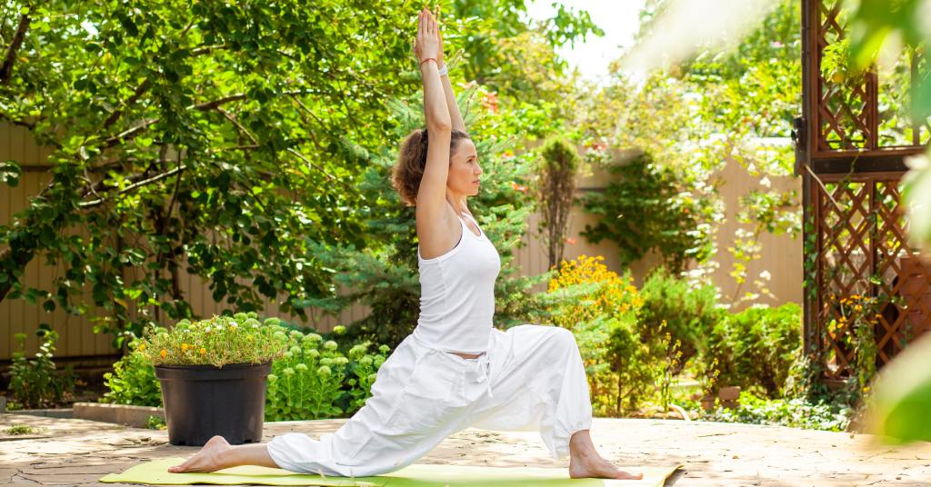 A young woman wearing a baggy white outfit practices yoga in a backyard space with a garden and natural elements.
