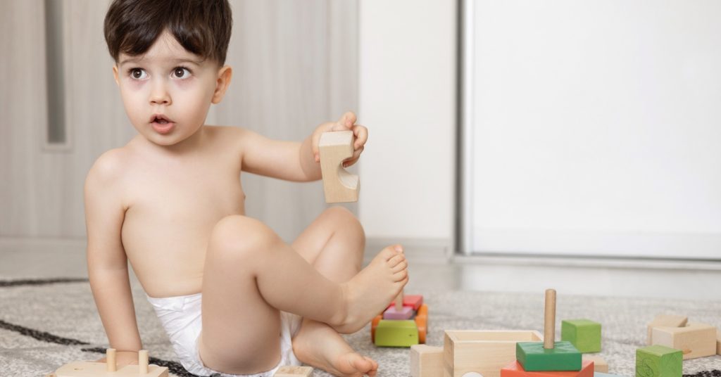 A young boy, dressed only in pull-ups, sitting on a rug and playing with wooden toy cars and blocks.