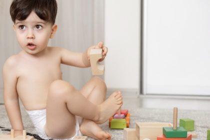 A young boy, dressed only in pull-ups, sitting on a rug and playing with wooden toy cars and blocks.
