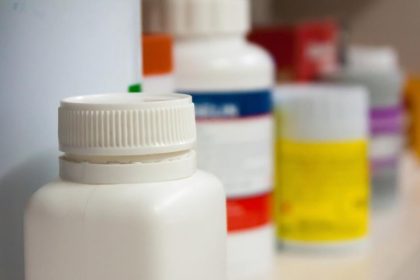 A close-up of a white pill bottle. There are multiple other bottles with labels blurred on the shelf behind the white bottle
