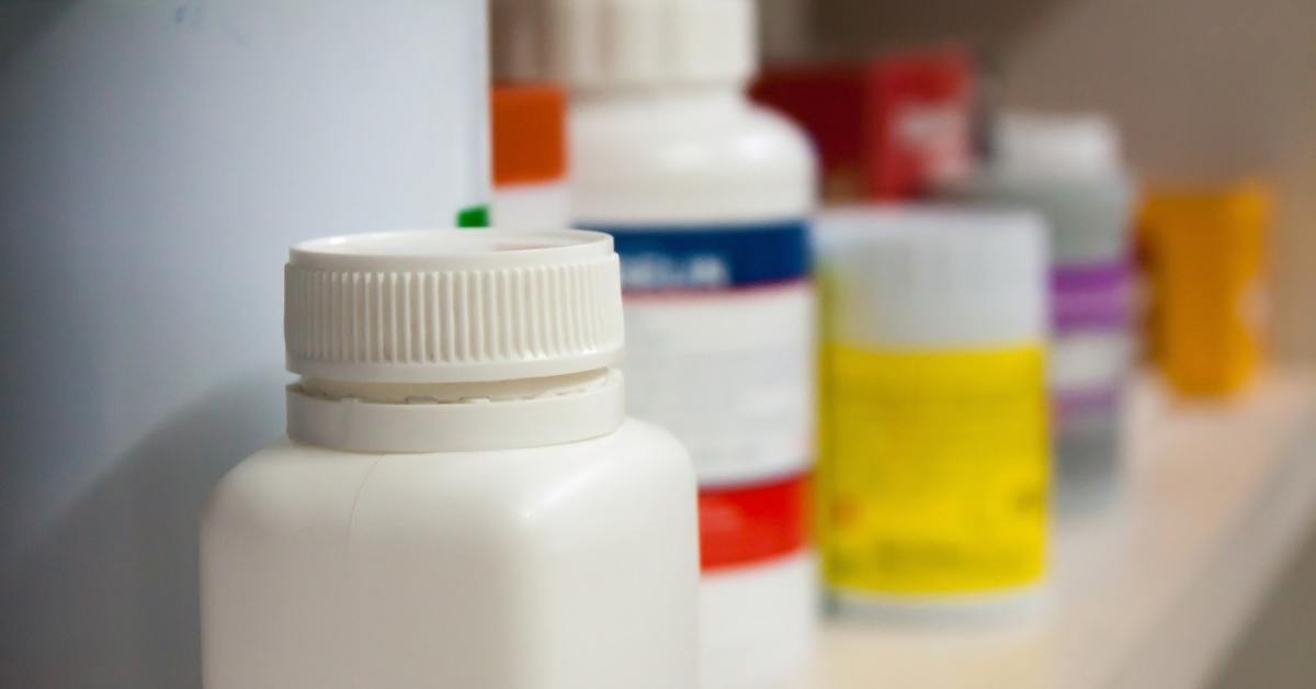 A close-up of a white pill bottle. There are multiple other bottles with labels blurred on the shelf behind the white bottle