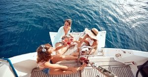 Three women sit on the back end of a white boat wearing summer clothing. They are raising their glasses for a toast.