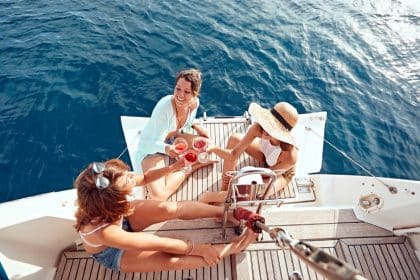 Three women sit on the back end of a white boat wearing summer clothing. They are raising their glasses for a toast.
