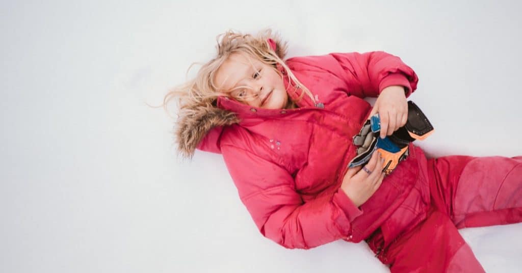 A young girl in a puffy, pink jacket laying on her back in the snow with her blonde hair messy.