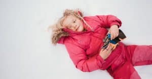 A young girl in a puffy, pink jacket laying on her back in the snow with her blonde hair messy.