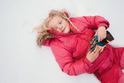A young girl in a puffy, pink jacket laying on her back in the snow with her blonde hair messy.