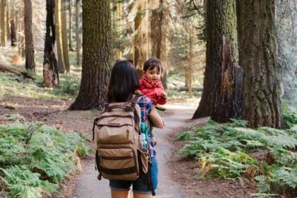 A young mother carries her daughter, who is a toddler, and a backpack, as she walks a trail in a forest.