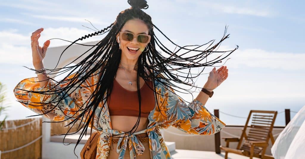 A woman with long box braids wears a colorful beach cover-up and sunglasses while smiling on a summer vacation.