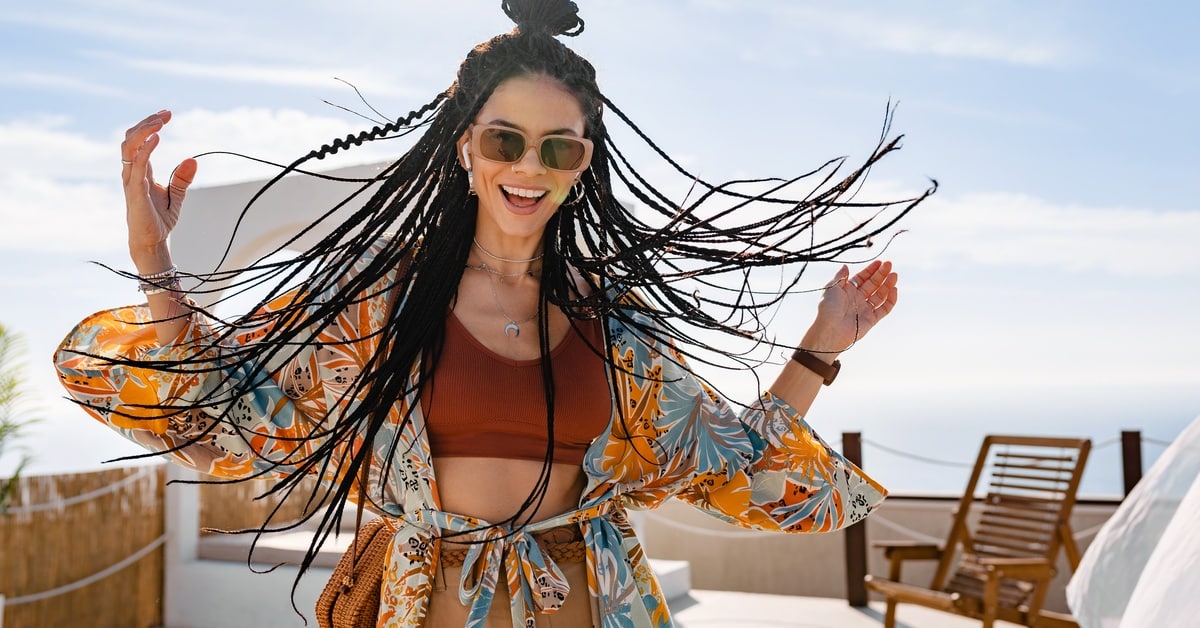 A woman with long box braids wears a colorful beach cover-up and sunglasses while smiling on a summer vacation.