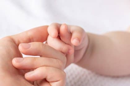 A close-up view shows a newborn gripping an adult's pointer finger. The background is a white blanket.