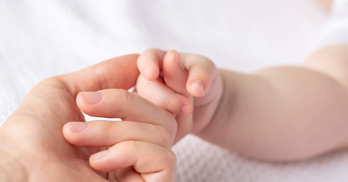 A close-up view shows a newborn gripping an adult's pointer finger. The background is a white blanket.